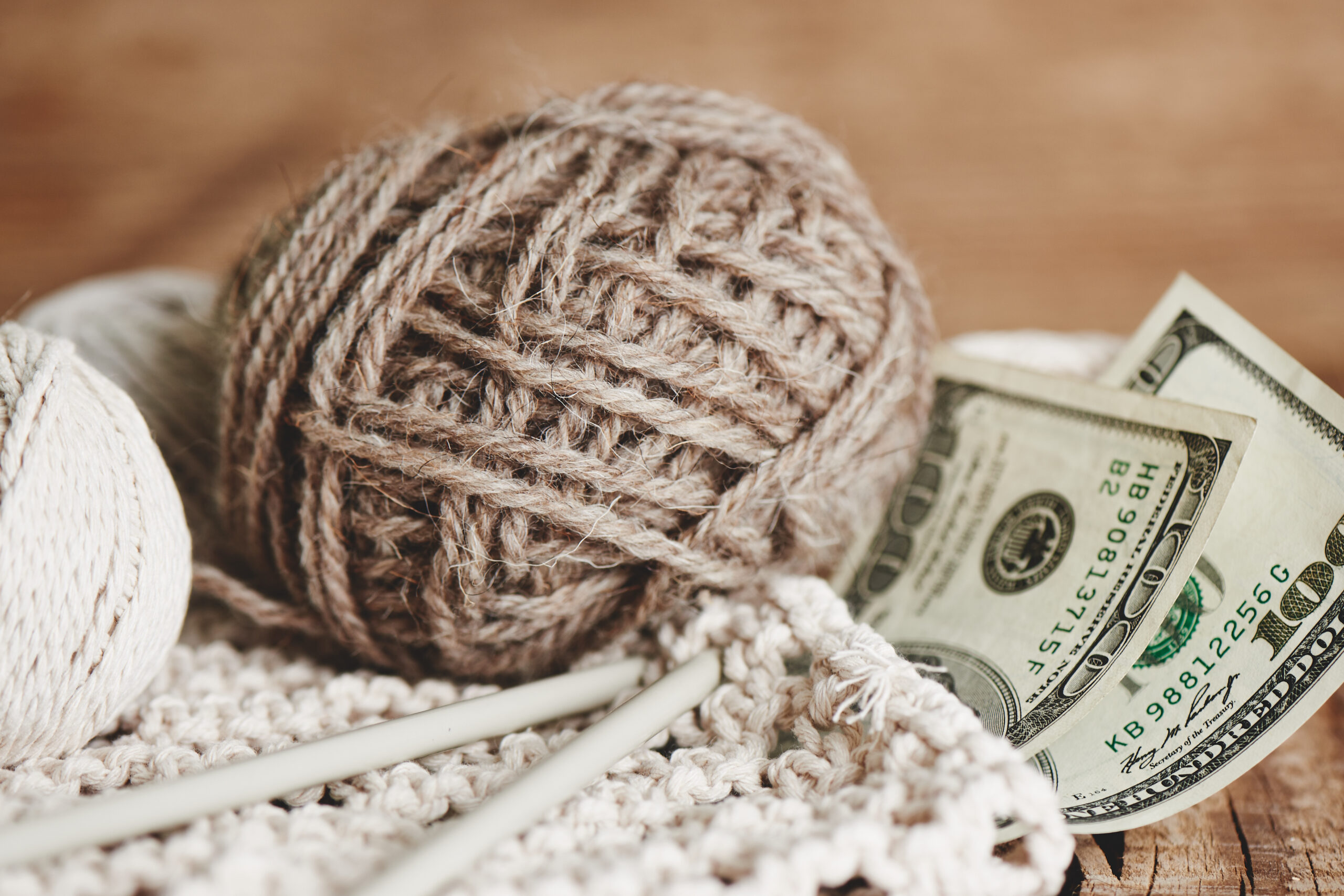 Profitable hobby. Earnings on needlework. Balls of natural color yarn, knitting needles and money on a wooden table.